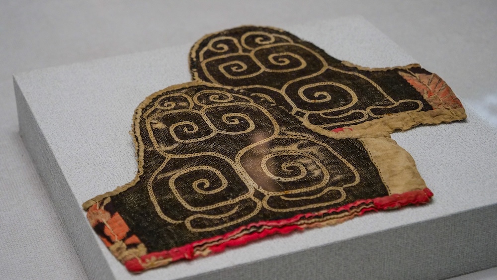 Historic Patterned Textile Exhibit at Hakodate Museum of Northern Peoples, Hokkaido, Japan A historic textile fragment displayed at the Hakodate Museum of Northern Peoples in Hokkaido, Japan, featuring intricate tan spiral patterns on dark fabric, with worn edges and sections of faded red stitching.