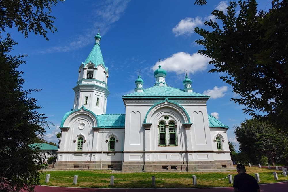The white walls, green copper roofs, small onion domes and bell tower of Hakodate Orthodox Church in Motomachi, Hakodate, Hokkaido, Japan, seen from across a lawn under a blue sky with scattered clouds.