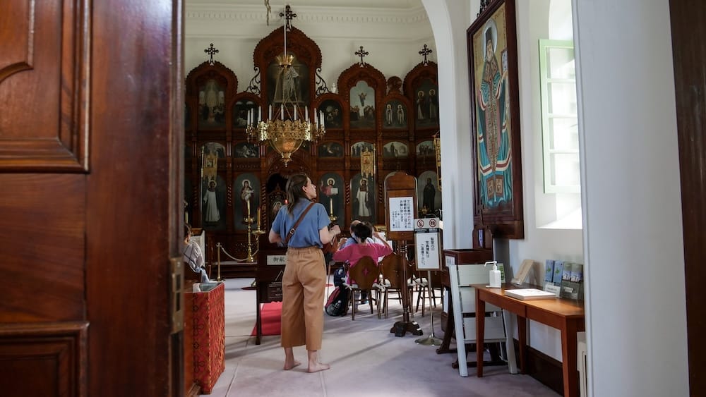 Audrey Bergner stands barefoot inside the Hakodate Orthodox Church in Motomachi, Hakodate, Hokkaido, Japan, facing an ornate gilded iconostasis with painted icons and a hanging candle chandelier as other visitors sit nearby.
