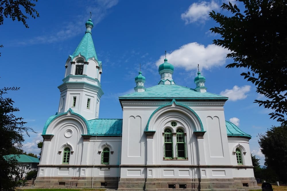 Hakodate Orthodox Church en Motomachi, Hakodate, Hokkaido, Japón, con paredes exteriores blancas, techos verde turquesa y distintivas torres ortodoxas enmarcadas por árboles bajo un cielo azul.