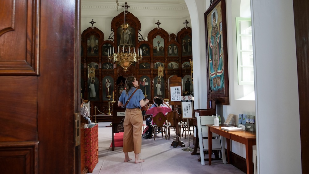 Hakodate Orthodox Church Interior in Motomachi, Hokkaido, Japan – Iconostasis and Audrey Bergner Visiting Audrey Bergner standing barefoot inside Hakodate Orthodox Church in Motomachi, Hokkaido, Japan, with an ornate wooden iconostasis, religious paintings, a hanging chandelier and visitors seated nearby.