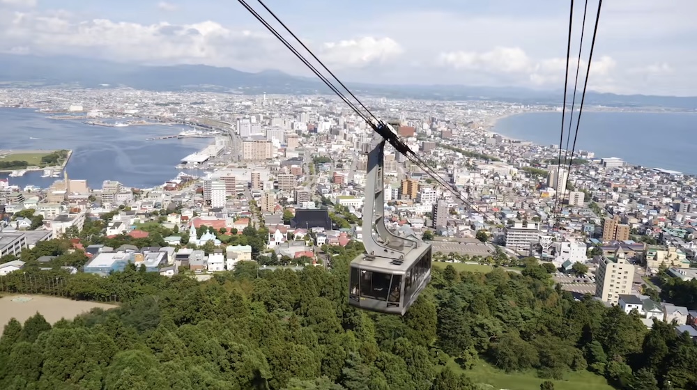 Hakodate Ropeway Cable Car Above Mount Hakodate Forest with Panoramic Views of the City and Both Bays, Hokkaido, Japan Hakodate Ropeway cable car travelling above the forested slopes of Mount Hakodate in Hokkaido, Japan, with the city stretching across a narrow peninsula between two bays and mountains on the horizon.
