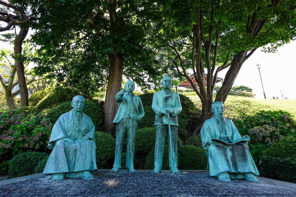 Four bronze statues of the Hakodate Shitenno in Motomachi Park, Hakodate, Hokkaido, Japan: Imai Ichiemon, Hirata Bun'emon, Watanabe Kumashiro and Hiratsuka Tokizo, arranged beneath leafy trees.