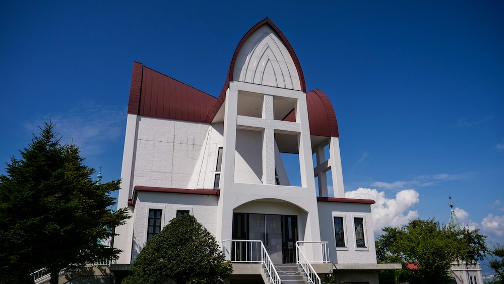 Hakodate St. John's Anglican Church, Hokkaido, Japan – Modern White Facade, Open Cross Tower and Curved Roof Angled exterior view of Hakodate St. John's Anglican Church in Motomachi, Hokkaido, Japan, showing its white walls, large open cross-shaped tower, curved reddish-brown roof, entrance stairs and surrounding trees.