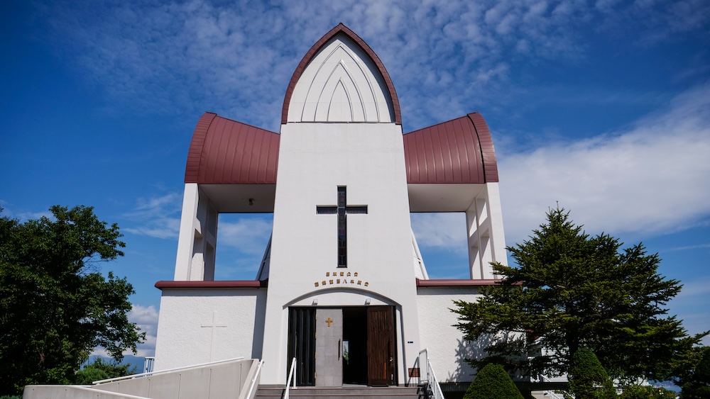 Hakodate St. John's Anglican Church in Motomachi, Hokkaido, Japan – Modern Exterior, Cross Window and Entrance Front view of Hakodate St. John's Anglican Church in Motomachi, Hakodate, Hokkaido, Japan, showing its white facade, large cross-shaped window, curved reddish-brown roof sections and entrance steps beneath a blue sky.