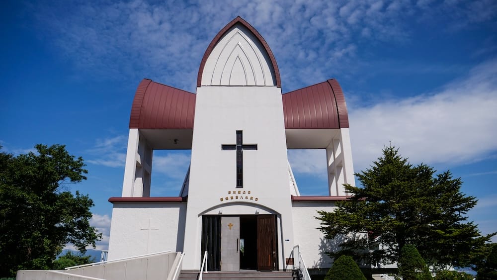The white facade of Hakodate St. John's Church in Motomachi, Hakodate, Hokkaido, Japan, with a pointed arch, curved maroon roof sections, a large dark cross and open doors under a bright blue sky.