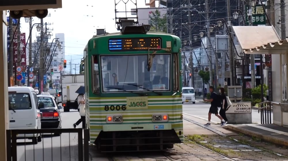 Tranvía de Hakodate cerca de Goryokaku Park en Hakodate, Hokkaido, Japón, sobre la red urbana Un tranvía verde de Hakodate detenido sobre vías a nivel de calle en Hakodate, Hokkaido, Japón, con peatones, autos, cables aéreos y un cartel cercano que señala hacia Goryokaku Park.