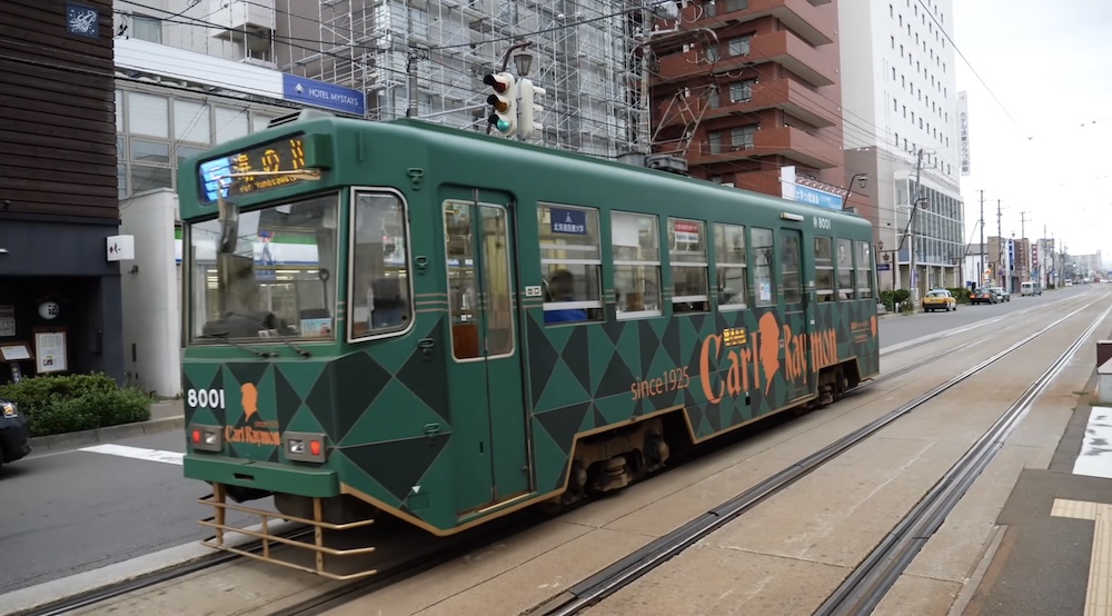 Hakodate Tram Line 5, Green Streetcar Number 8001, Public Transport in Hakodate, Hokkaido, Japan Green Hakodate city tram number 8001 operating on Line 5 along street-level railway tracks in Hakodate, Hokkaido, Japan, with commercial buildings, overhead wires and an intersection visible around it.
