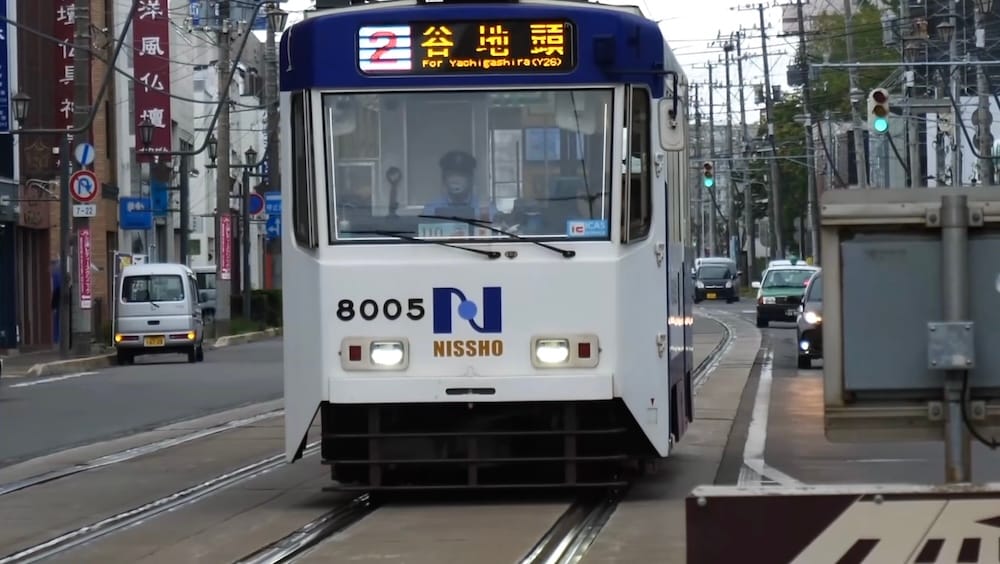 Tranvía de la Ruta 2 de Hakodate con destino a Yachigashira sobre vías urbanas en Hakodate, Hokkaido, Japón Tranvía blanco de la Ruta 2 de Hakodate circulando por vías urbanas en Hokkaido, Japón, con el indicador frontal iluminado mostrando Yachigashira como destino y tránsito detrás.