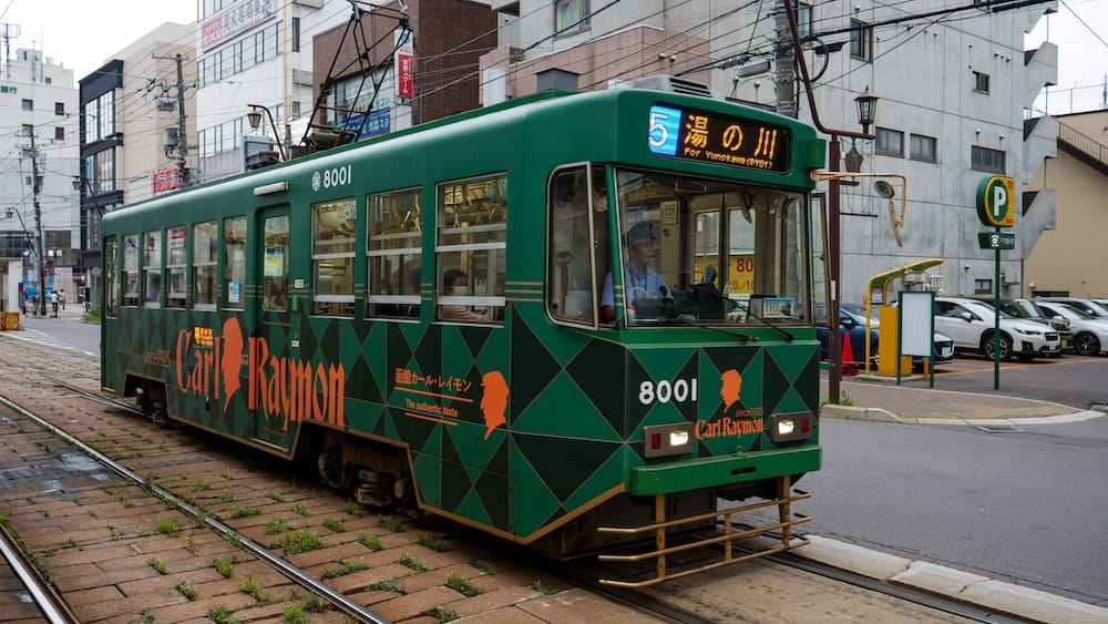 Green Hakodate tram on Route 5 travelling through Hakodate, Hokkaido, Japan, with the destination display showing Yunokawa and overhead power lines running above the city street.