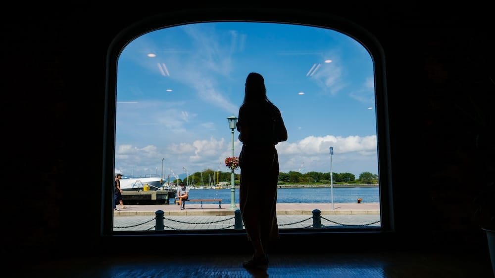 Audrey Bergner stands in silhouette before a large arched window overlooking the Hakodate waterfront in Hakodate, Hokkaido, Japan, with the harbour, promenade, lamp post and blue sky visible outside.