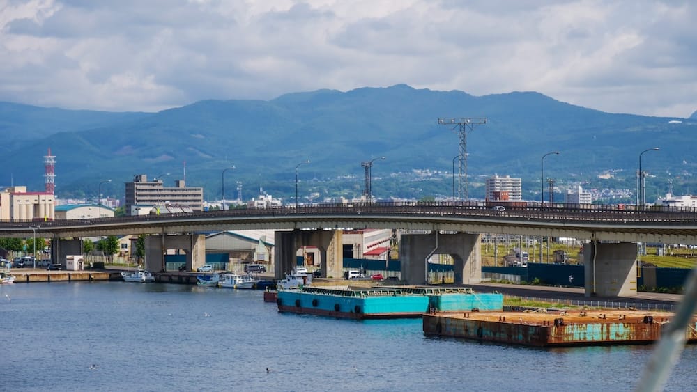Hakodate waterfront harbour view with boats, bridge, city and mountains in Hokkaido, Japan Ground-level waterfront view in Hakodate, Hokkaido, Japan, showing harbour water, moored boats, an elevated bridge, city buildings and a broad mountain ridge beneath cloudy skies.