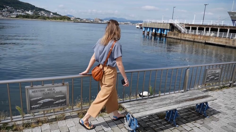 Waterfront walk, Hakodate, Hokkaido, Japan – Audrey Bergner Audrey Bergner walks along a waterfront railing in Hakodate, Hokkaido, Japan, overlooking the harbour, a ferry dock and distant hills across the water.