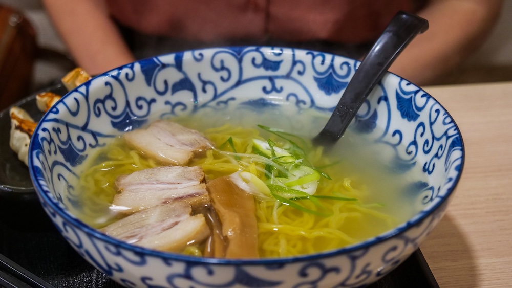 Steaming close-up of salt ramen in a blue-and-white bowl, with yellow noodles in a clear golden broth under three slices of pork, a strip of bamboo shoot and chopped green onion, in Hakodate, Hokkaido, Japan.