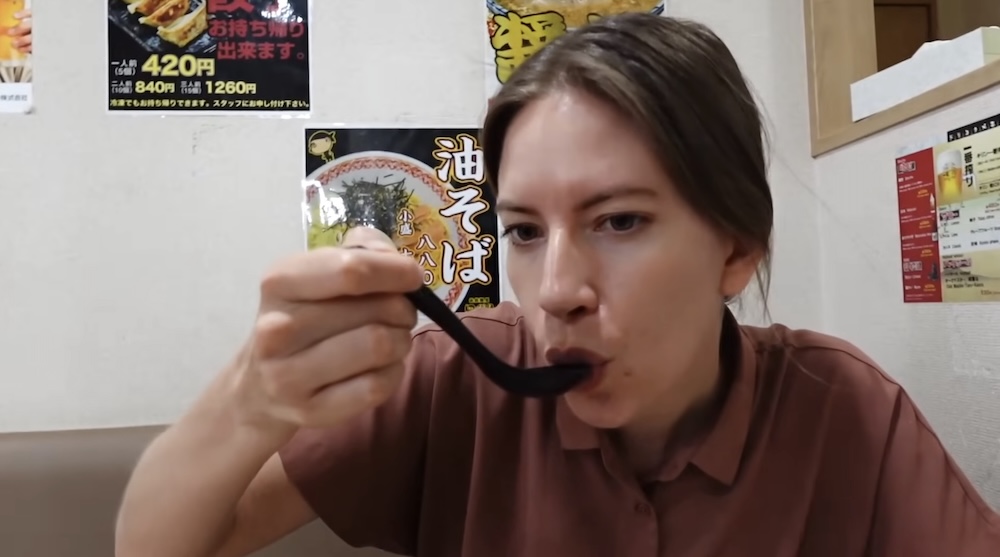 Audrey Bergner in a rust-coloured shirt sips broth from a black spoon at a booth in a ramen shop in Hakodate, Hokkaido, Japan, with gyoza and ramen posters on the wall behind her.