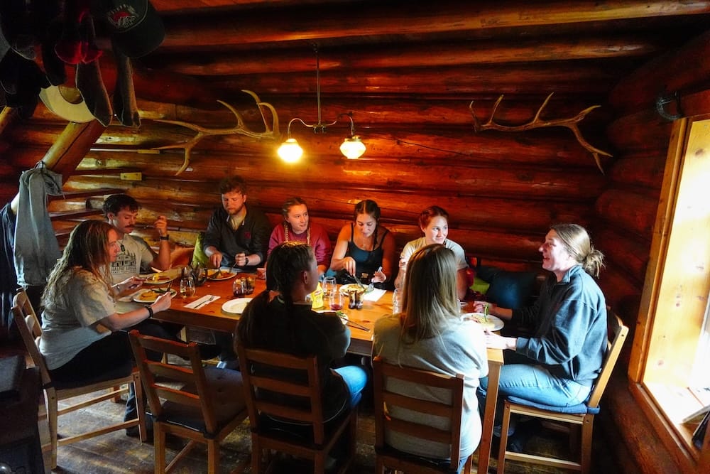 Cena comunal en Halfway Lodge, Parque Nacional Banff, Alberta, Canadá, durante la travesía a caballo Un grupo comparte una comida alrededor de una mesa comunal dentro de Halfway Lodge, en el Parque Nacional Banff, Alberta, Canadá, en un interior de troncos con astas en las paredes y luz entrando por una ventana.