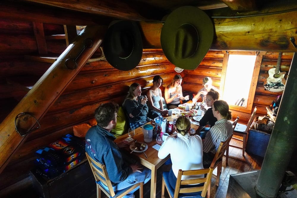 Comedor comunitario de Halfway Lodge en el Parque Nacional Banff, Alberta, Canadá, durante una travesía a caballo por el backcountry Huéspedes reunidos alrededor de una mesa de madera dentro de Halfway Lodge, en el Parque Nacional Banff, Alberta, Canadá, con paredes de troncos, sombreros vaqueros, estufa a leña y espacio común compacto.