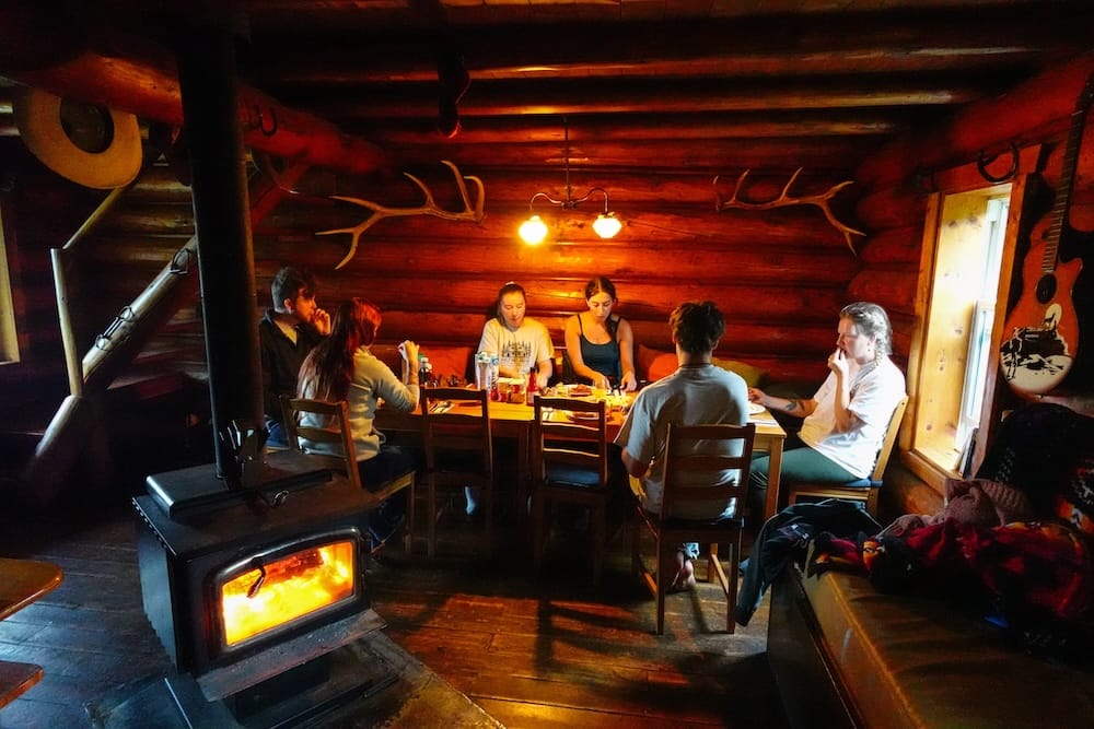 Halfway Lodge dining room in Banff National Park, Alberta, Canada with wood stove and guests during the horseback trip Guests sit around a dinner table beside a glowing wood stove inside Halfway Lodge in Banff National Park, Alberta, Canada, showing the warm communal atmosphere that contrasted with the rugged backcountry riding outside.