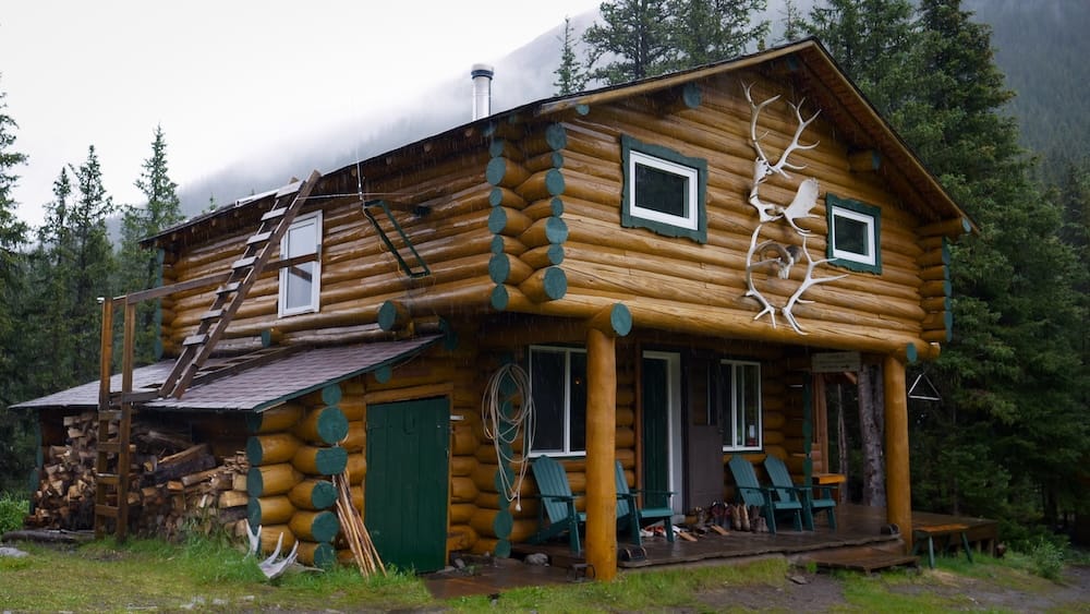 Exterior histórico de Halfway Lodge en el Parque Nacional Banff, Alberta, Canadá, mostrando la cabaña de troncos de dos pisos Exterior de troncos de dos pisos de Halfway Lodge, Parque Nacional Banff, Alberta, Canadá, con astas en la fachada, sillas en el porche, leña apilada y bosque con neblina alrededor.