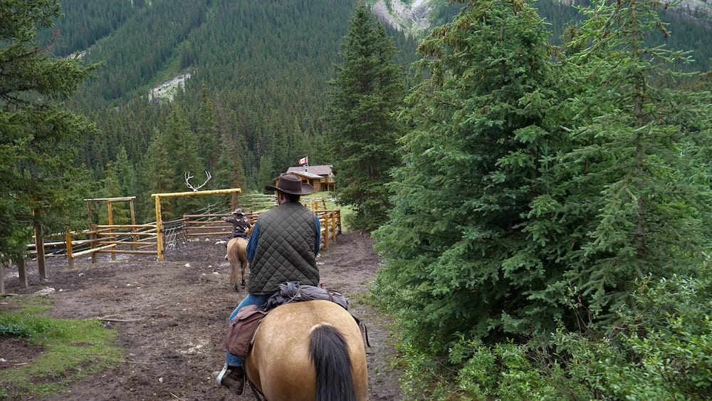 Horseback riders arrive at Halfway Lodge in Banff National Park, Alberta, Canada, passing wooden corrals and evergreen forest on a muddy backcountry trail.