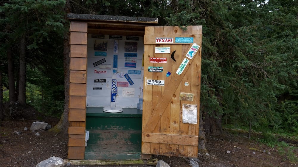 Letrina de Halfway Lodge en el Parque Nacional Banff, Alberta, Canadá, con estructura rústica de madera en el bosque Letrina de Halfway Lodge en el Parque Nacional Banff, Alberta, Canadá, con una estructura de madera gastada, puerta abierta, stickers humorísticos y un denso bosque de coníferas alrededor.