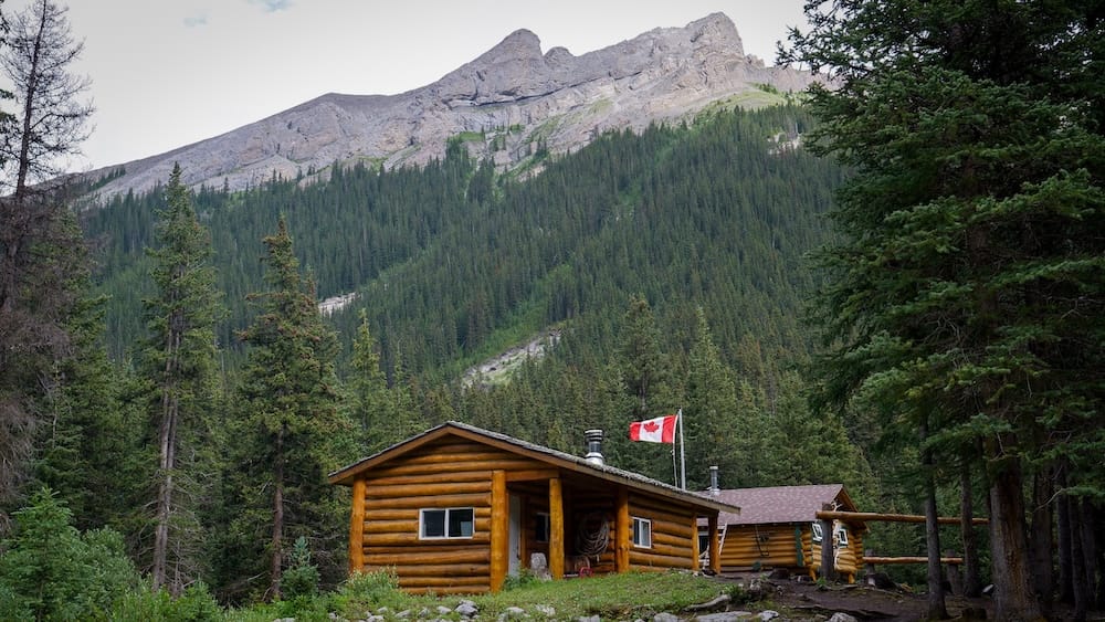 Halfway Lodge in Banff National Park, Alberta, Canada with rustic log cabins beneath a forested mountain ridge Halfway Lodge sits beneath a steep forested mountain ridge in Banff National Park, Alberta, Canada, with rustic log cabins, evergreen trees and a Canadian flag marking the remote backcountry setting.