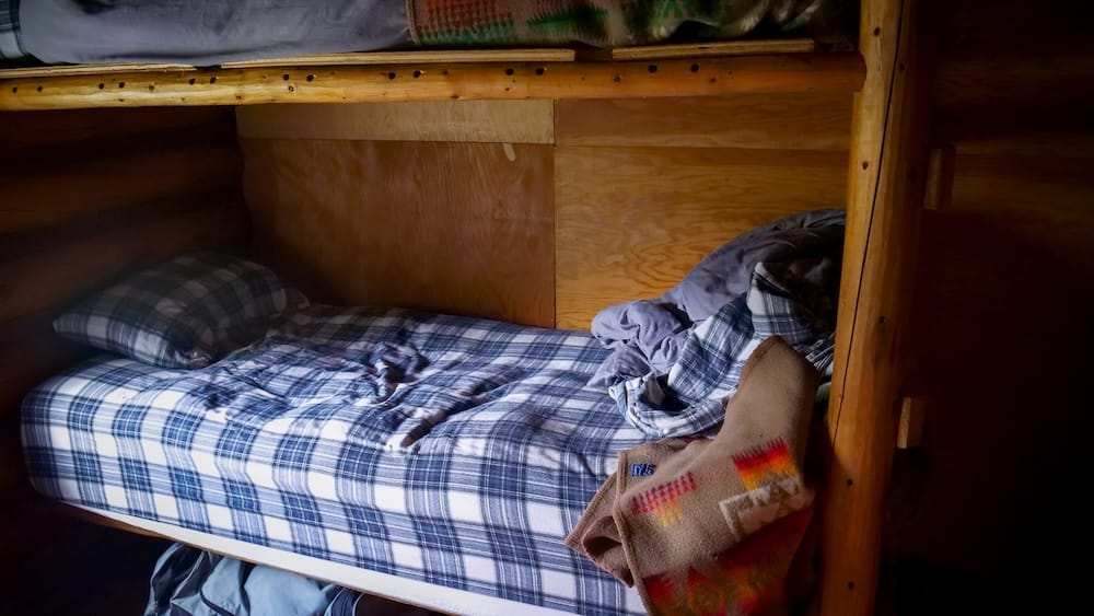 Habitación con cuchetas en Halfway Lodge, Parque Nacional Banff, Alberta, Canadá, con alojamiento compartido rústico Cama cucheta rústica dentro de una habitación compartida de Halfway Lodge, Parque Nacional Banff, Alberta, Canadá, con ropa de cama a cuadros, mantas y equipo de cabalgata alrededor.