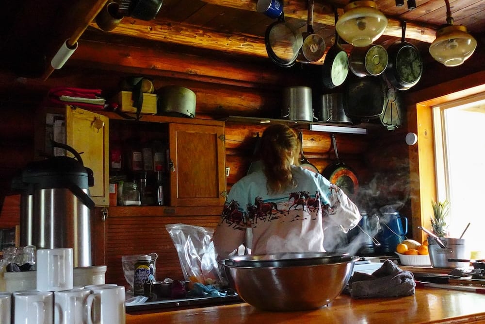 Cocina rústica de Halfway Lodge en el Parque Nacional Banff, Alberta, Canadá, con comida preparándose para los huéspedes Una persona prepara comida en la cocina rústica de Halfway Lodge, Parque Nacional Banff, Alberta, Canadá, rodeada de sartenes colgadas, alacenas sencillas de madera y ollas humeantes.