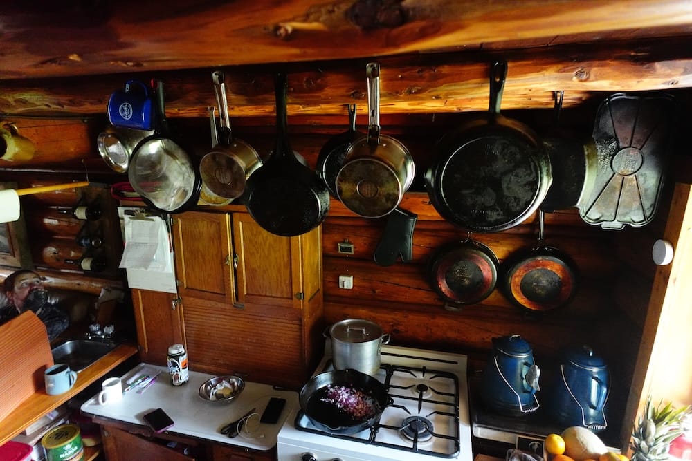 Cocina rústica de Halfway Lodge en el Parque Nacional Banff, Alberta, Canadá, con utensilios colgados e interior de troncos Cocina rústica dentro de Halfway Lodge, en el Parque Nacional Banff, Alberta, Canadá, con sartenes colgadas, cocina a gas, pavas, paredes de madera y un espacio compacto para cocinar en la cabaña.