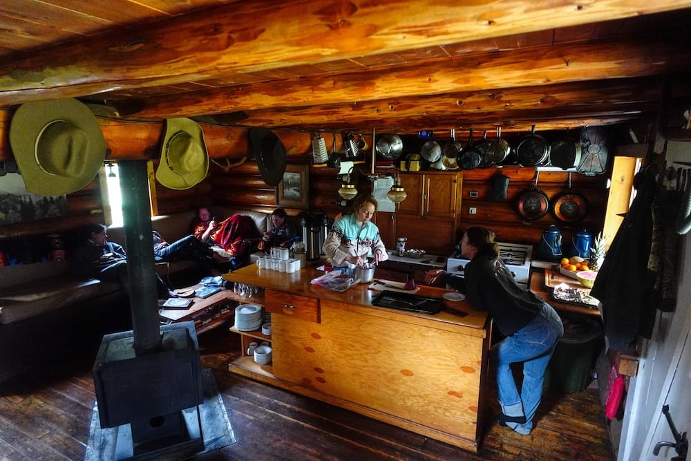 Cocina comunitaria de Halfway Lodge en el Parque Nacional Banff, Alberta, Canadá, con estufa a leña e interior rústico Acogedora cocina y sala comunitaria dentro de Halfway Lodge, Parque Nacional Banff, Alberta, Canadá, con huéspedes descansando alrededor de una estufa a leña mientras se prepara comida bajo utensilios colgados.