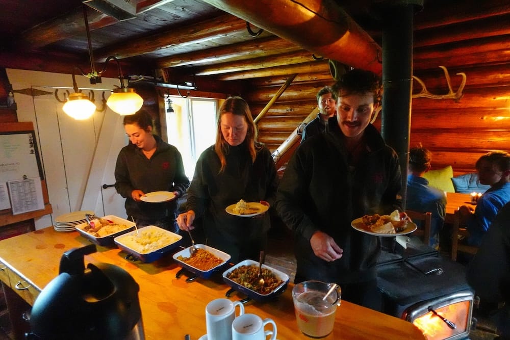 Cena para servirse con porciones generosas en Halfway Lodge durante una travesía a caballo de Banff Trail Riders Los huéspedes se sirven la cena de fuentes compartidas dentro de Halfway Lodge, en el Parque Nacional Banff, Alberta, Canadá, llenando sus platos junto a la estufa a leña durante una travesía de Banff Trail Riders.