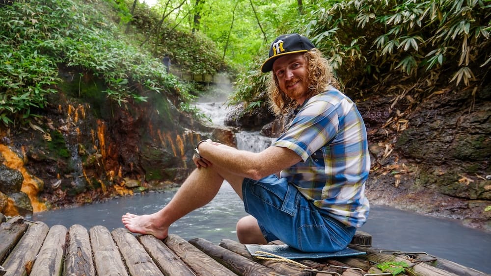Samuel Jeffery, a barefoot traveller sits beside a steaming outdoor hot spring in Noboribetsu, Hokkaido, wearing a black Hanshin Tigers baseball cap during a trip through Japan.
