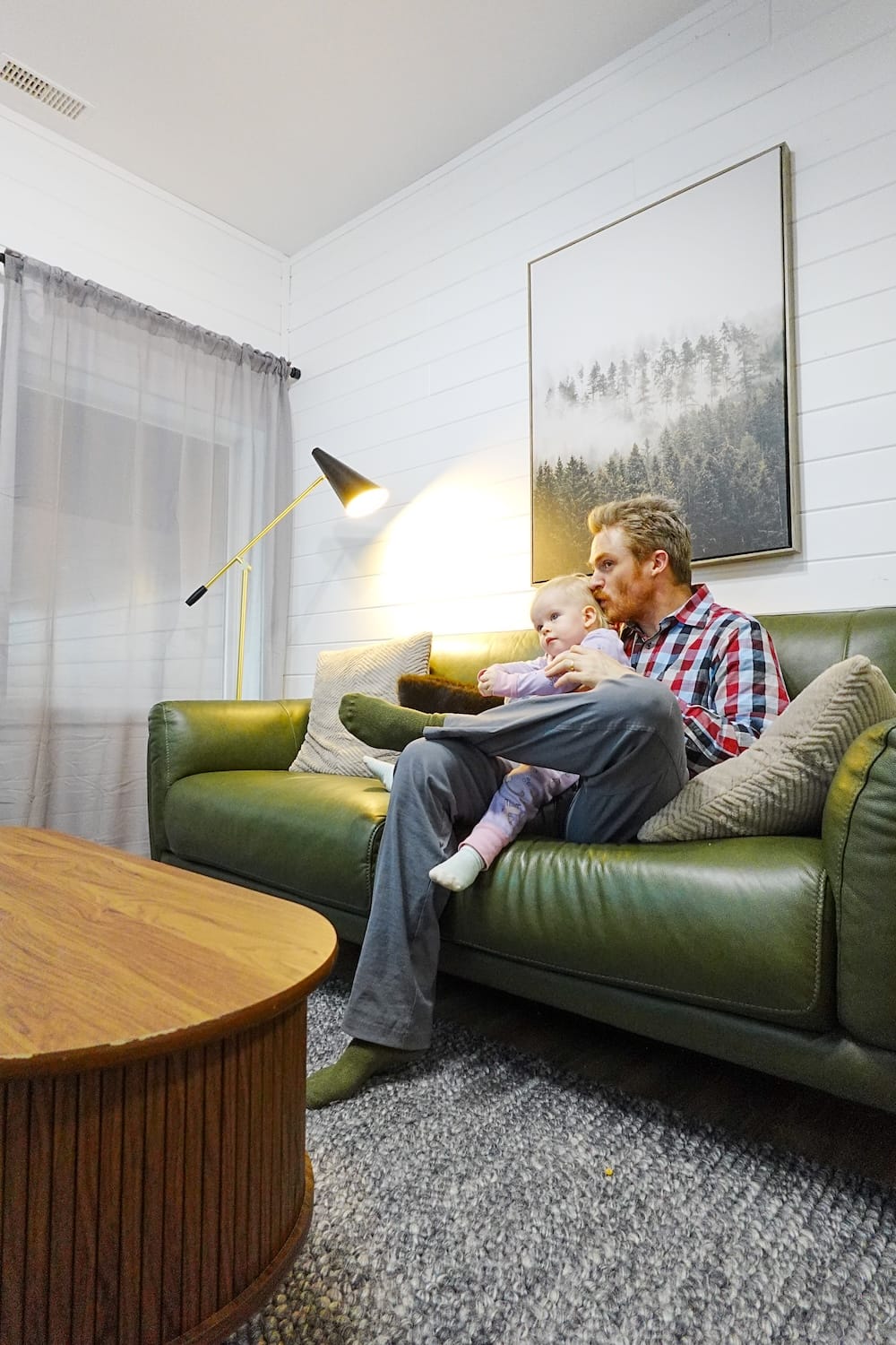Samuel Jeffery sitting with Aurelia Jeffery on a green sofa inside family accommodation near Banff, Alberta, Canada, with white panelled walls, a floor lamp and mountain-themed artwork behind them.