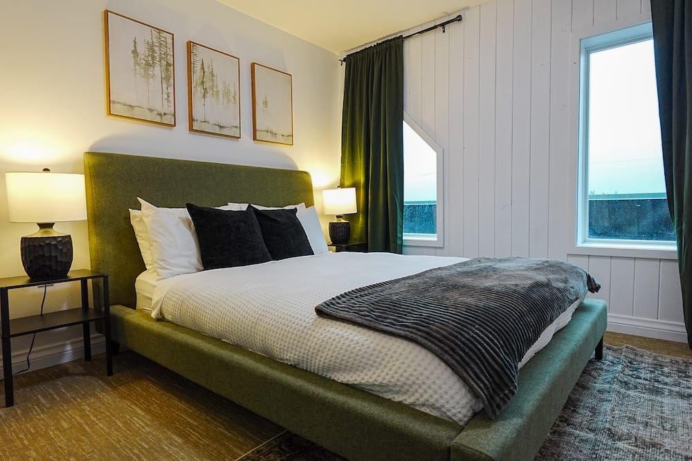 A modern accommodation bedroom in Harvie Heights, Alberta, Canada, with a large bed, green upholstered headboard, bedside lamps, forest artwork, dark curtains and tall windows.