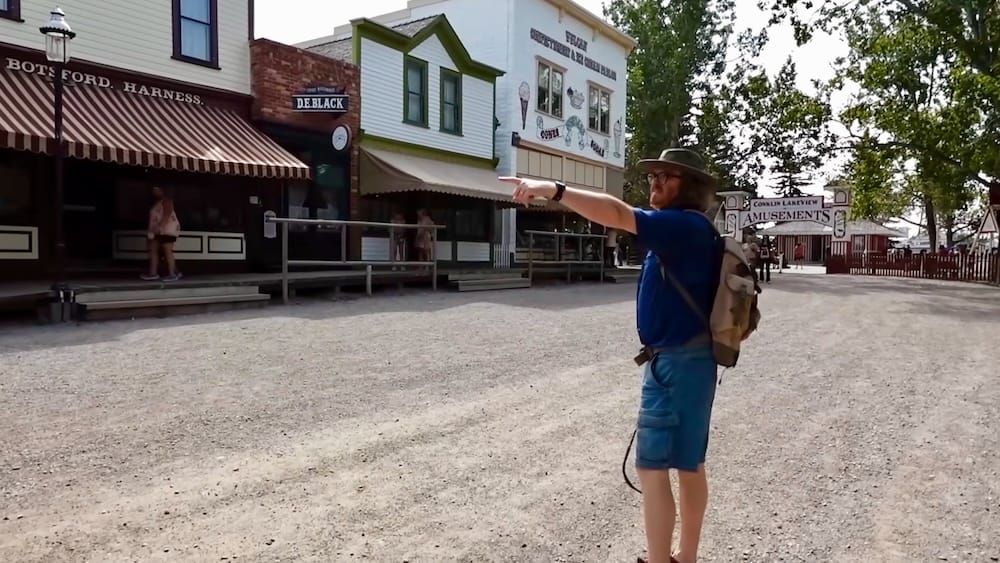 Samuel Jeffery walking through the historic streets of Heritage Park Historical Village in Calgary, Alberta, Canada, with period storefronts and the Antique Midway entrance in the background.