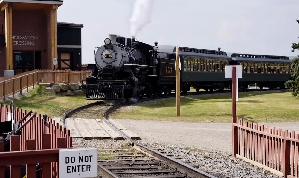 Steam locomotive pulling vintage passenger cars along the railway tracks at Heritage Park Historical Village in Calgary, Alberta, Canada, with the train rounding a curve through the grounds.