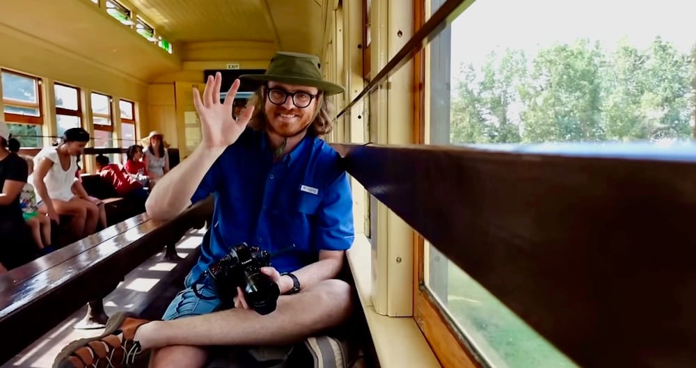 Samuel Jeffery seated inside a vintage passenger car on the Heritage Park steam train in Calgary, Alberta, Canada, waving while holding a camera as other riders sit farther back.