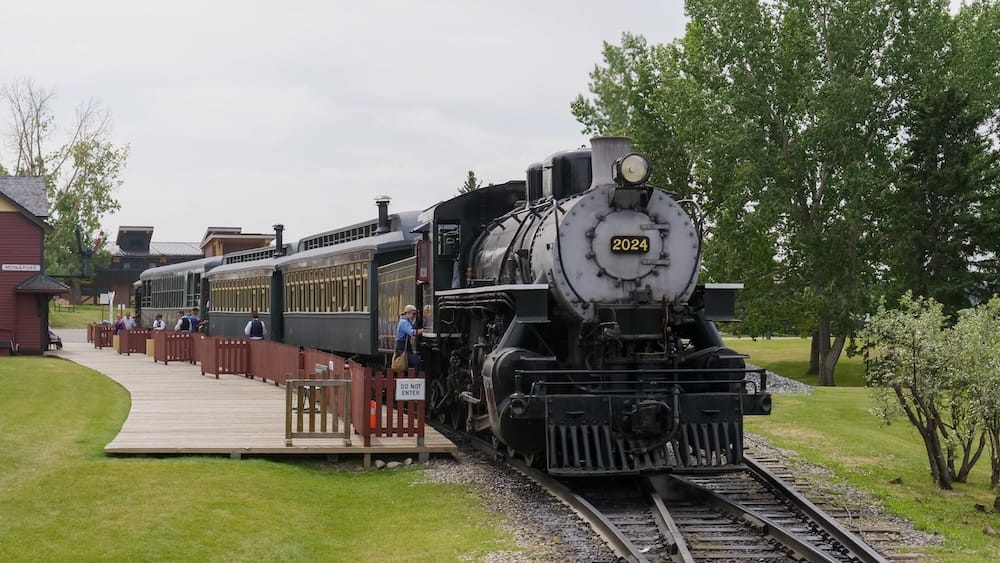 Vintage steam locomotive and passenger cars stopped at Heritage Park Historical Village in Calgary, Alberta, Canada, beside a wooden platform with trees and historic buildings in the background.