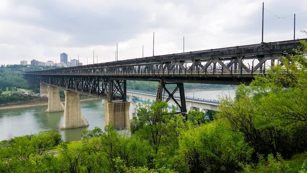 Edmonton High Level Bridge spanning the North Saskatchewan River valley in Alberta, Canada, with its steel structure, concrete piers, green riverbanks and city buildings visible in the distance.