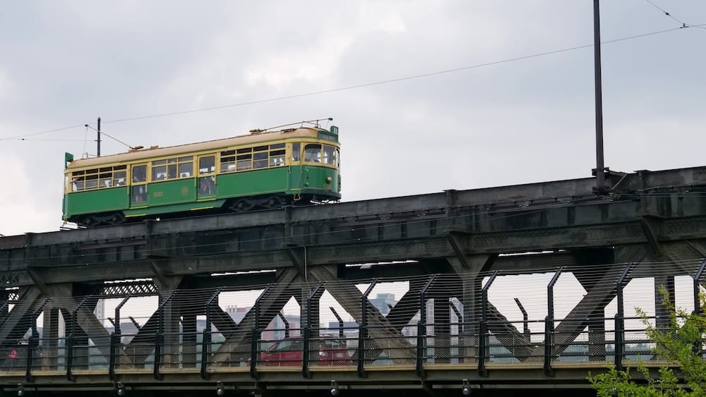 A heritage green and cream streetcar crosses the steel truss span of the High Level Bridge in Edmonton, Alberta, Canada, with the city skyline visible through the girders below.