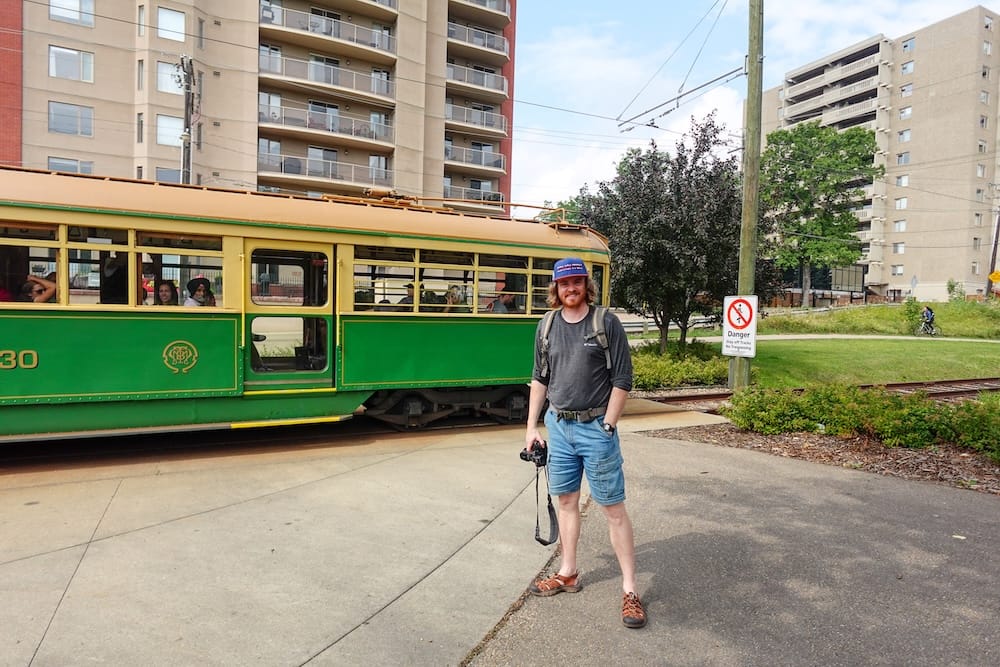 Samuel Jeffery stands with a camera beside Edmonton’s green-and-cream High Level Bridge Streetcar, with tracks, overhead wires and apartment buildings visible in Edmonton, Alberta, Canada.