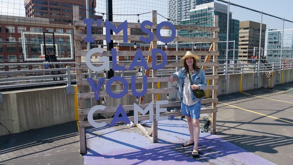 Audrey Bergner beside the I’m So Glad You Came sign at High Park in Calgary, Alberta, Canada Audrey Bergner stands beside the “I’m So Glad You Came” sign at High Park in Calgary, Alberta, Canada, with rooftop structures and downtown buildings visible behind her.