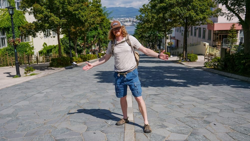 Samuel Jeffery smiles with arms outstretched on a wide stone-paved hillside street in Hakodate, Hokkaido, Japan, with trees, buildings and the harbour and mountains visible at the bottom of the slope on a sunny day.