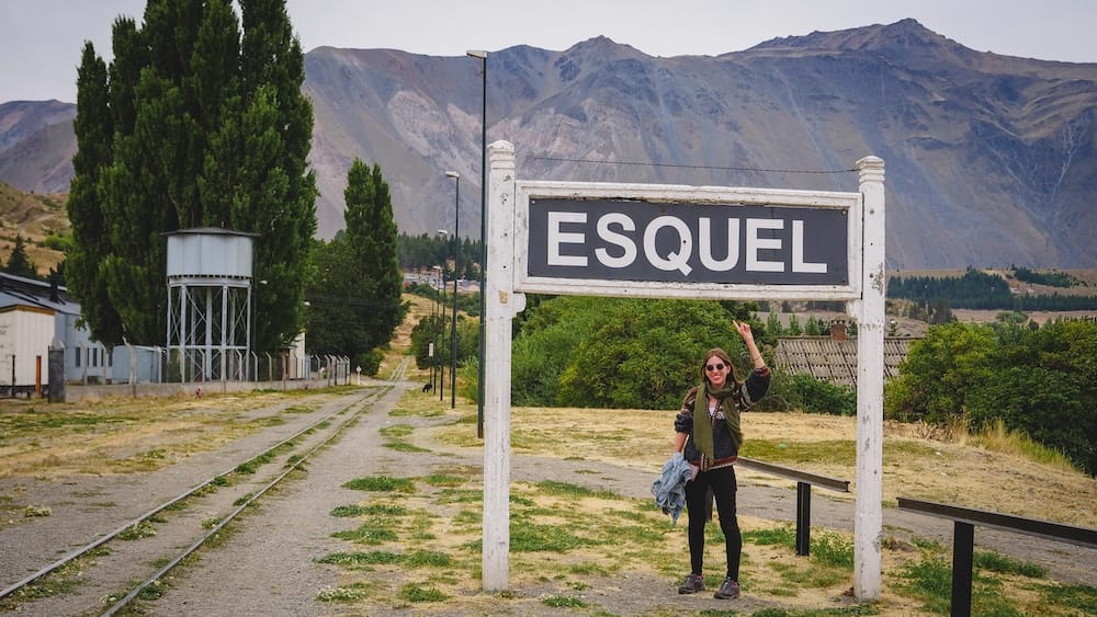 Audrey Bergner stands beneath the large Esquel station sign at the historic railway grounds in Esquel, Chubut, Patagonia, Argentina, with narrow-gauge tracks, poplar trees and rugged Andean peaks behind her.