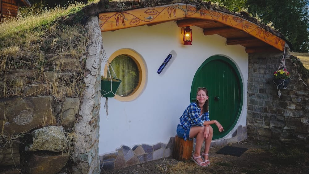 Hobbit-style earth-sheltered cabin, El Bolsón, Río Negro, Argentine Patagonia — Audrey Bergner Audrey Bergner sitting on a wooden stump outside a whimsical earth-sheltered hobbit house with a round green door in El Bolsón, Río Negro, Argentine Patagonia.