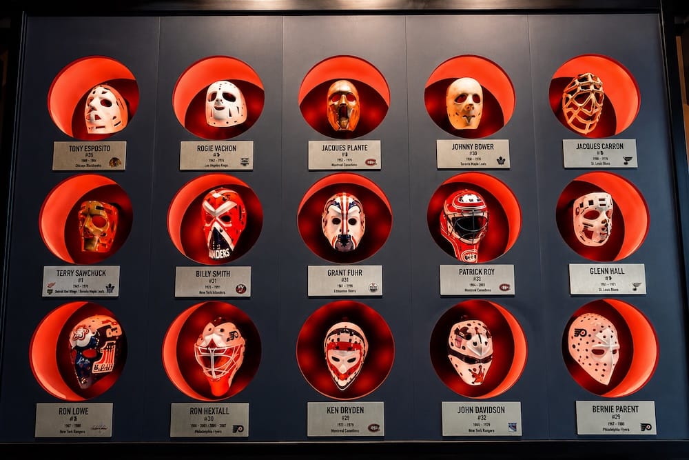 Historic hockey goalie mask display near Rogers Place in Edmonton ICE District, Alberta, Canada A wall display of vintage hockey goalie masks near Rogers Place in Edmonton’s ICE District, Alberta, Canada, with historic masks arranged in illuminated red niches and labelled by player.