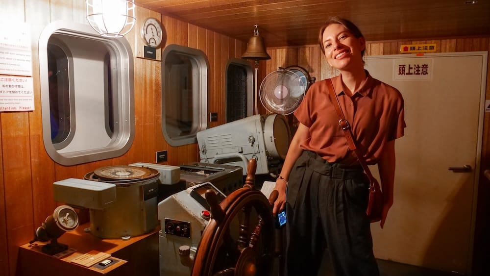Audrey Bergner stands beside the wheel and controls of the fishing-boat simulator at the Hokuyo Museum in Hakodate, Hokkaido, Japan, inside a wood-panelled mock wheelhouse with maritime equipment.