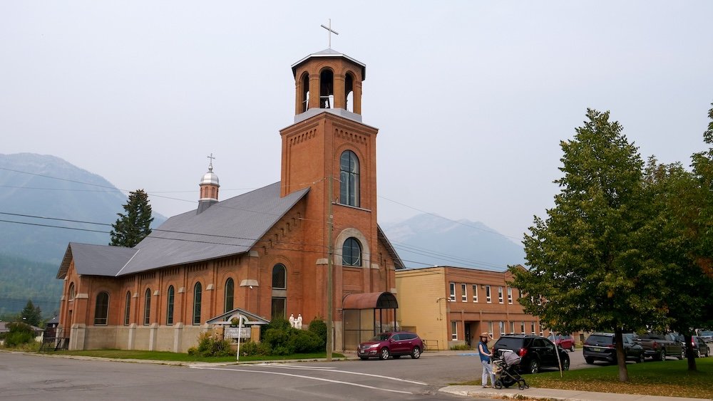 Holy Family Catholic Church in Fernie, British Columbia, a red-brick heritage church with a tall bell tower and arched windows, photographed during a family stroll with Audrey Bergner and baby Aurelia in a stroller near downtown streets and mountain backdrops.
