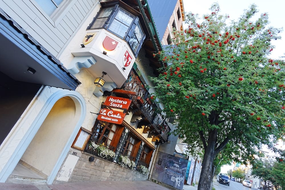 Hostería Suiza in downtown Bariloche, Río Negro, Patagonia, Argentina, with alpine-style architecture Hostería Suiza stands on a downtown Bariloche street beneath a berry-laden tree, with alpine-style details, carved wood balconies and visible hotel signage in Río Negro, Patagonia, Argentina.