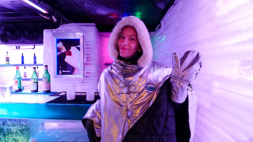 Audrey Bergner wearing a silver insulated suit inside an ice bar in El Calafate, Santa Cruz, Patagonia, Argentina, surrounded by sculpted ice walls, neon lighting, bottles and a frozen counter.